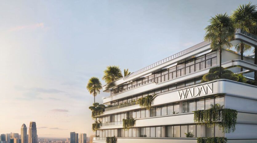 Un bâtiment blanc moderne avec de grandes fenêtres et des terrasses verdoyantes, portant l'inscription "WADANI", se détache sur la ligne d'horizon de la ville au coucher du soleil - un exemple élégant de la vie urbaine à Tresora by Wadan, avec de grands gratte-ciel en arrière-plan.