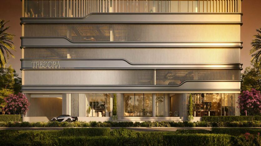 Un bâtiment moderne à l'architecture métallique et linéaire et aux grandes baies vitrées, étiqueté "Tresora by Wadan", est entouré de verdure, de palmiers et de buissons fleuris, avec une voiture garée à l'entrée pendant le coucher du soleil.