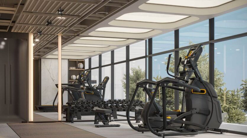 Salle de sport moderne à Tresora by Wadan avec de grandes fenêtres, comprenant des machines elliptiques, un rack de poids avec des haltères, un banc d'entraînement, et la lumière naturelle entrant à l'intérieur ; la verdure est visible à l'extérieur.