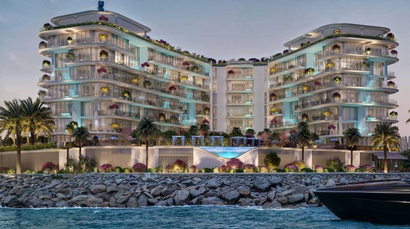 La luxueuse et moderne Villa Del Garda, située en bord de mer, se compose de deux ailes incurvées, de balcons en verre, de palmiers, de fleurs colorées et d'une piscine centrale éclairée. Elle est vue de l'eau, avec une digue en pierre devant et un bateau partiellement visible sur la droite.