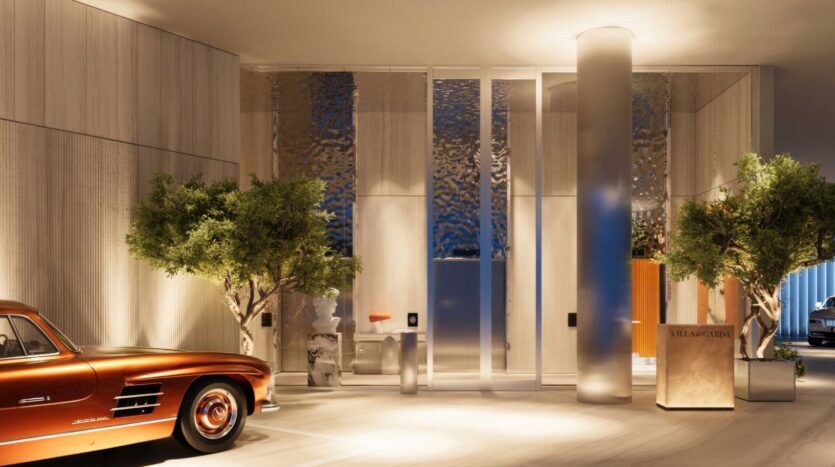 Un hall d'entrée luxueux et moderne avec de hautes portes vitrées, des arbres verdoyants, un éclairage chaleureux et une voiture vintage brillante de couleur cuivre garée à l'intérieur. Un petit bureau de réception indique "VILLA ARTEFACTO" à la Villa Del Garda.