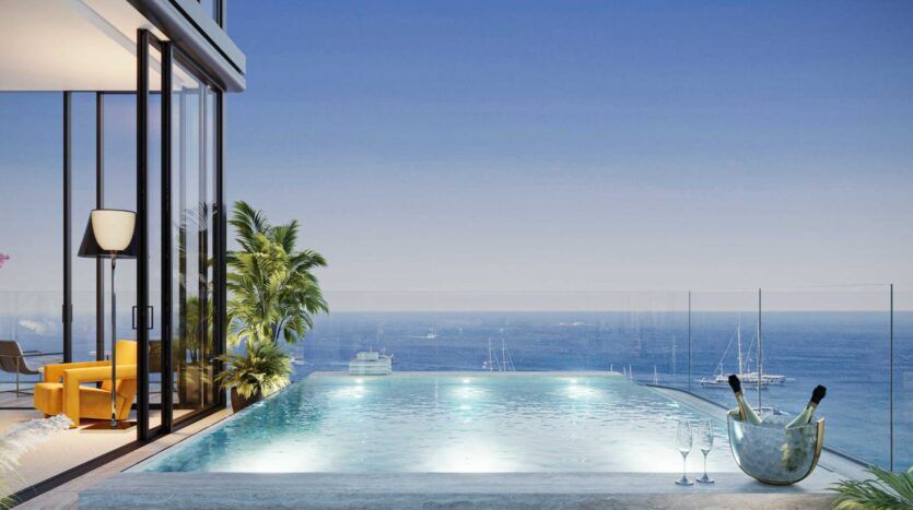Piscine à débordement surplombant l'océan au coucher du soleil à la Villa Del Garda, avec des chaises longues et une salle moderne aux murs de verre sur la gauche. Du champagne dans un seau à glace et deux coupes sont posés au bord de la piscine, tandis que des voiliers dérivent sur l'eau calme et bleue.
