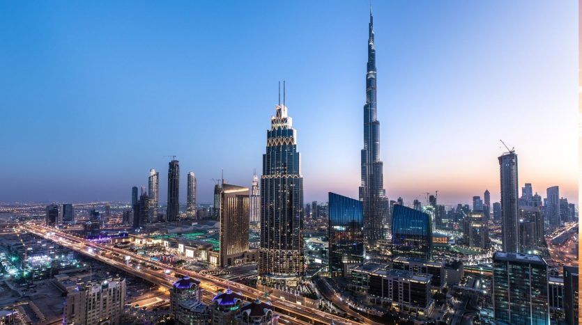 Une vue panoramique des meilleurs quartiers de Dubaï au crépuscule mettant en valeur l'horizon illuminé avec le Burj Khalifa et d'autres gratte-ciel sur un ciel bleu décoloré.