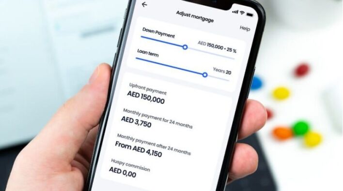 Main d'une personne tenant un smartphone affichant une application de calcul d'hypothèques de crédit immobilier Dubaï, avec des champs pour l'acompte, la durée du prêt et les mensualités, visibles à côté de trombones colorés.