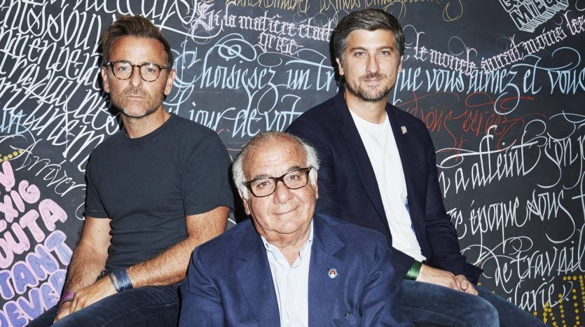 Trois hommes assis devant un tableau noir couvert d'écritures colorées de style graffiti dans une villa de Dubaï, regardant la caméra. Le plus jeune est au milieu et tous sont habillés en tenue professionnelle décontractée.