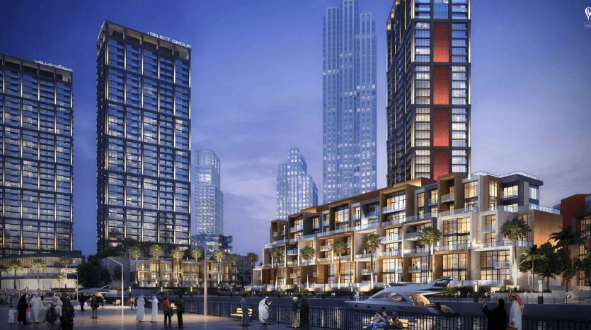 Vue nocturne du Peninsula Dubai, un développement luxueux en bord de mer avec des gratte-ciel modernes, une promenade remplie de monde et un yacht amarré près d'élégants bâtiments bas.