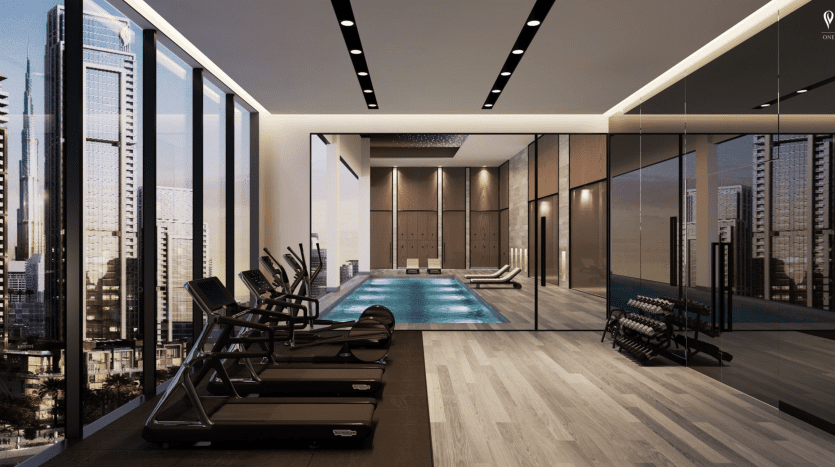 Une salle de sport luxueuse en hauteur avec de grandes fenêtres donnant sur les toits de la ville au crépuscule, gérée par Peninsula Select Group Dubai. L'intérieur comprend des tapis roulants, des poids libres et une piscine intérieure éclairée.