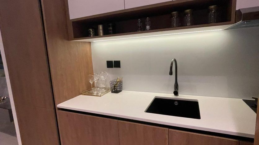 Une kitchenette moderne dans Prime Residency 3 comprend des armoires en bois élégantes, un évier noir intégré et un comptoir blanc. La verrerie est exposée sur l'étagère et l'éclairage ambiant améliore l'ambiance.