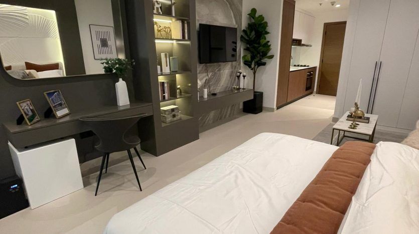 Chambre au design moderne dans Prime Residency 3 comprenant un grand lit avec une literie blanche et en terre cuite, des étagères intégrées décorées et une télévision. Un couloir mène à une autre pièce.