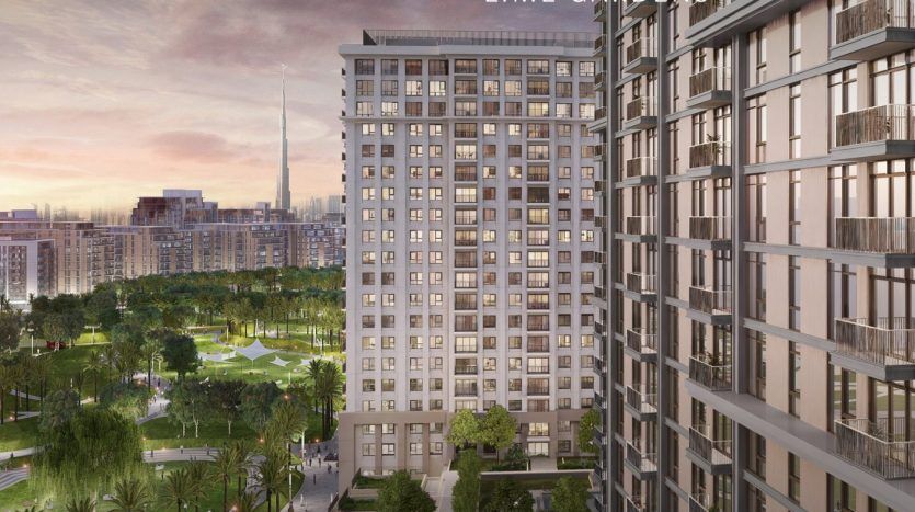 Immeuble résidentiel de grande hauteur avec une façade moderne, surplombant un parc luxuriant et d'autres bâtiments urbains au coucher du soleil, avec vue sur un grand gratte-ciel de Dubaï.