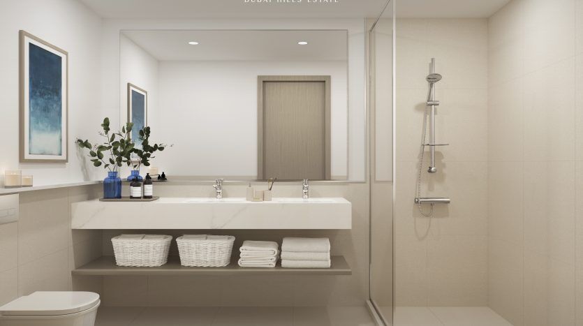 Intérieur de salle de bains moderne avec douche en verre, vanité blanche surmontée de bouteilles et de fleurs bleues, paniers en osier et serviettes soigneusement pliées. Des œuvres d'art sont suspendues au-dessus des toilettes. Le texte indique "Hills Park Dubai Hills