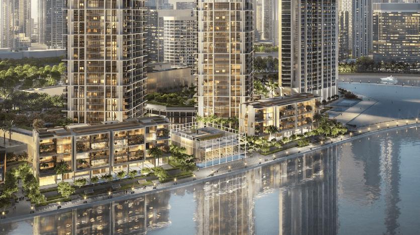 Vue aérienne d'un développement moderne en bord de mer avec plusieurs appartements de grande hauteur, des espaces verts luxuriants et des façades éclairées au crépuscule dans un cadre urbain à Dubaï.