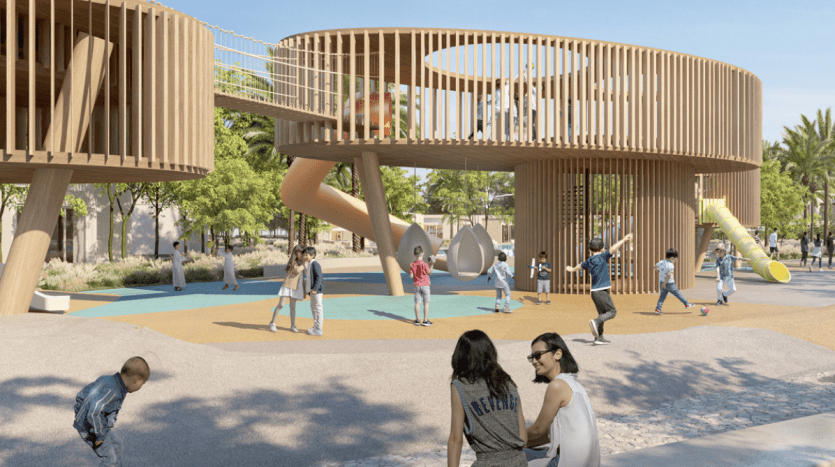 A lively playground featuring a large circular wooden structure with slides and climbing areas. Children play on the ground while adults sit nearby, watching from comfortable benches amid sunny surroundings with lush green trees typical of