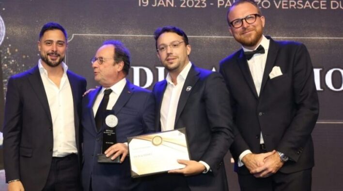 Quatre hommes en tenue de soirée, l'un titulaire d'un certificat et l'autre d'une médaille, souriant lors d'une cérémonie de remise de prix liée à l'investissement Dubaï. Détails de l'événement et date visibles en arrière-plan.