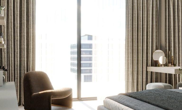 Une chambre moderne dans un appartement de Dubaï comprenant un lit luxueux, une chaise moelleuse, de grands rideaux et une baie vitrée donnant sur des gratte-ciel urbains. La lumière du soleil illumine le décor sophistiqué et élégant