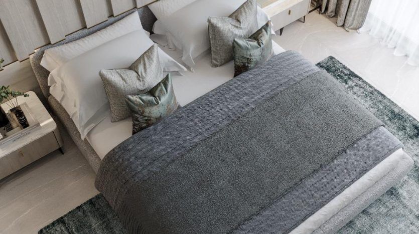 Une chambre moderne et minimaliste comprenant un grand lit avec des draps blancs et une couverture grise, un tapis bleu sarcelle en dessous et un plafonnier circulaire unique. Des tons neutres doux et une lumière naturelle créent une ambiance sereine dans cette villa
