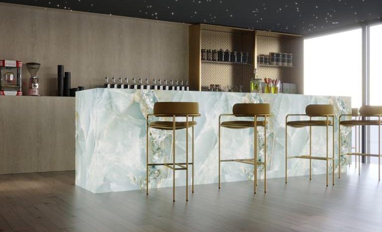 Un bar moderne éclairé par un plafond étoilé, doté d'un comptoir translucide en forme de glace avec des détails dorés et des tabourets hauts. Des étagères remplies de bouteilles sont visibles dans un intérieur en bois rappelant un appartement haut de gamme