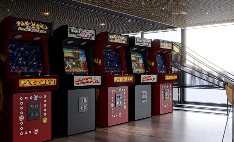 Une salle d'arcade moderne présentant une gamme de machines d'arcade classiques Pac-man sous un plafond doré et réfléchissant, avec du parquet et un coin salon éloigné dans une villa exclusive de Dubaï.