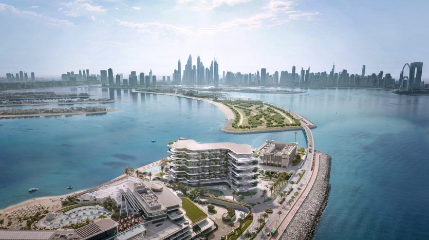 Vue aérienne d'un développement côtier moderne à Dubaï avec des bâtiments à plusieurs niveaux au design unique surplombant un lagon bleu, avec un horizon urbain en arrière-plan lointain sous un ciel clair.