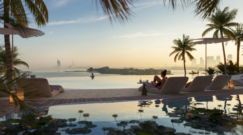 Une scène sereine au bord de la piscine au coucher du soleil avec une silhouette d'un paysage urbain de Dubaï au loin. Des feuilles de palmier encadrent le dessus, des chaises longues sont au premier plan et une personne se détend au bord de l'eau.
