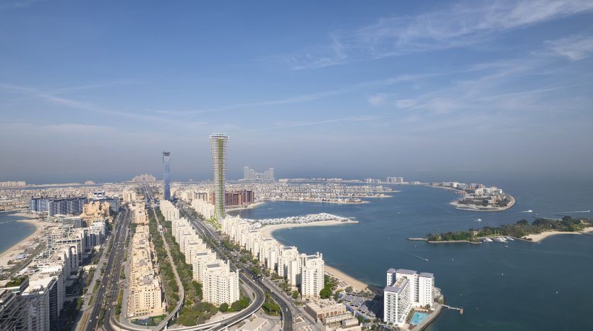 Vue aérienne d'une ville côtière avec des bâtiments modernes denses, notamment des gratte-ciel et des villas, entourée d'eaux bleu clair et de rivages incurvés, sous un ciel clair.