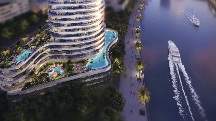 Vue aérienne d'un immeuble de grande hauteur moderne et curviligne avec des balcons éclairés, une piscine et une verdure luxuriante à Dubaï, à côté d'une rivière avec un yacht naviguant au crépuscule.