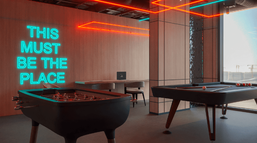 Salle de loisirs moderne avec des néons affichant "This must be the place", comprenant un baby-foot, une table de billard et un bureau avec un ordinateur dans un appartement de Dubaï.