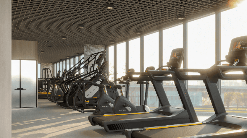 Salle de sport moderne avec des rangées de tapis roulants et de machines elliptiques faisant face à de grandes fenêtres offrant une vue sur le lever du soleil à Dubaï, créant une atmosphère d'entraînement lumineuse et accueillante.
