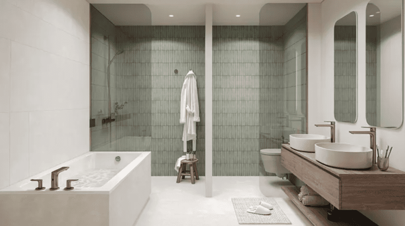 Salle de bains moderne comprenant une grande baignoire avec un rebord en bois et des luminaires en laiton, une douche à l'italienne avec des carreaux verts Verdes, des toilettes murales et une double vasque avec vasques, robinets en laiton et deux miroirs allongés. Un peignoir blanc est suspendu à côté de la douche.