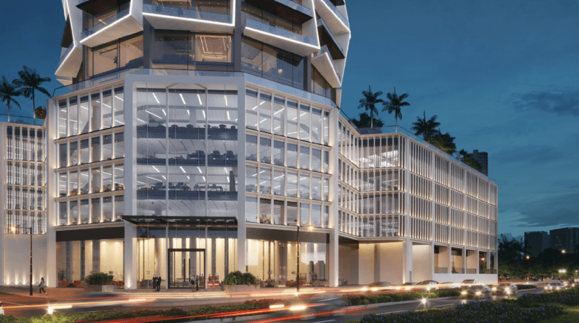 Un immeuble de bureaux moderne en verre est éclairé au crépuscule, avec une architecture géométrique, de grandes fenêtres révélant des intérieurs éclairés, des palmiers sur les terrasses et des phares de voiture flous sur la route devant.