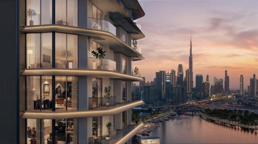 Une tour moderne avec des balcons en verre met en valeur l'art architectural en surplombant un port de plaisance et une rivière au coucher du soleil, avec la ligne d'horizon de la ville et le Burj Khalifa visibles en arrière-plan. Des lumières chaudes brillent à l'intérieur des appartements.