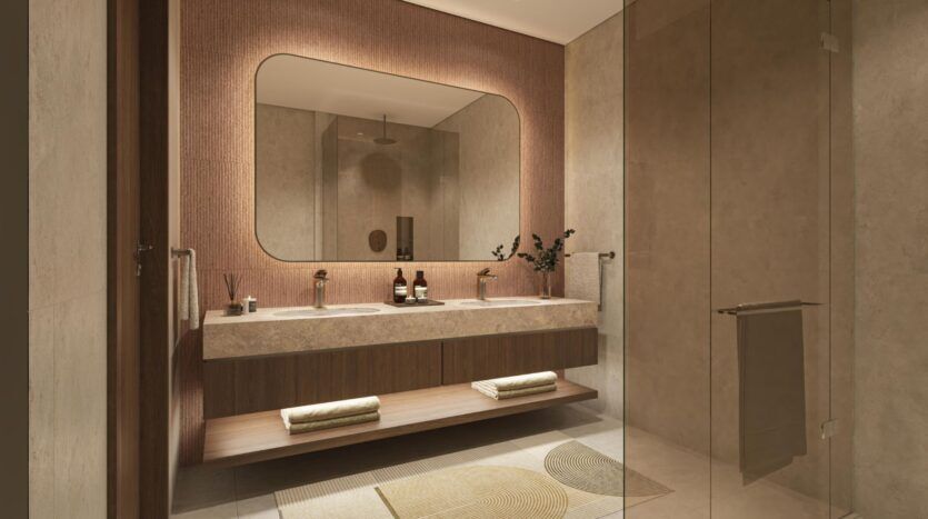 Une salle de bains moderne qui met en valeur l'art avec un grand miroir, un double lavabo, des finitions en bois brun et en pierre, des serviettes pliées sur une étagère, une cabine de douche en verre et un tapis décoratif sur un sol en carreaux de faïence clairs.