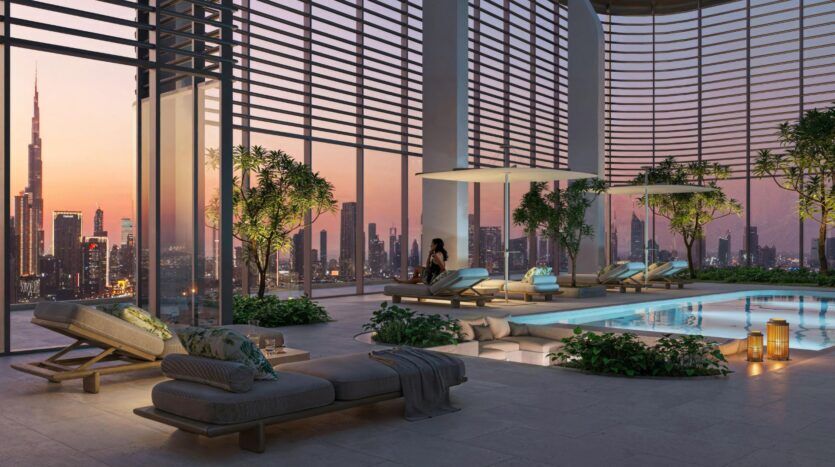 La luxueuse piscine intérieure, dotée de chaises longues, d'un décor moderne et de touches de verdure, met en valeur l'art, tandis que de grandes fenêtres encadrent une ligne d'horizon époustouflante de la ville au coucher du soleil ; une femme se détend sur une chaise près de la piscine scintillante.