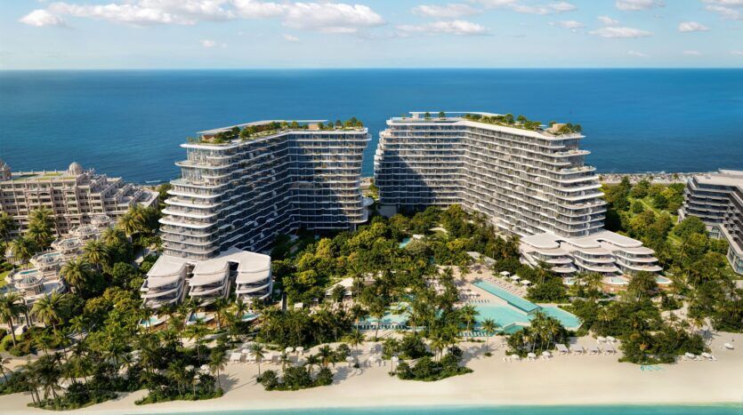 Vue aérienne des bâtiments modernes et incurvés de Bella by Passo, situés en bord de mer et entourés d'une végétation luxuriante, de piscines et d'espaces de détente. Ils sont parfaitement situés le long d'une plage de sable et d'un océan turquoise, sous un ciel bleu parsemé de nuages.