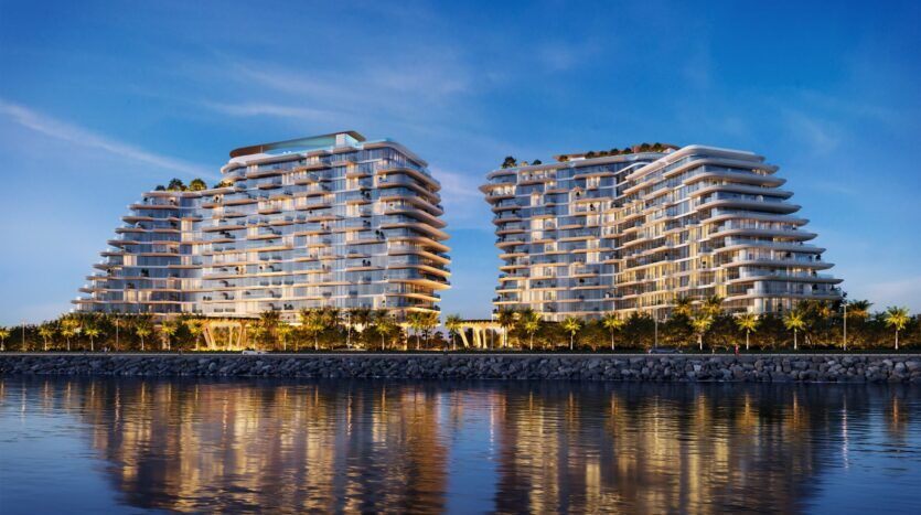 À Bella by Passo, deux immeubles résidentiels modernes de plusieurs étages avec des balcons incurvés se dressent côte à côte, illuminés au crépuscule et se reflétant dans le calme du front de mer, entouré de palmiers.