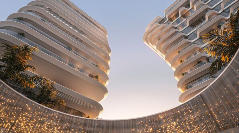 Vue en contre-plongée de deux bâtiments modernes, blancs et incurvés avec des balcons à Bella by Passo, des palmiers et une structure circulaire en contrebas ornée de lumières scintillantes, le tout dans un ciel clair au coucher du soleil.