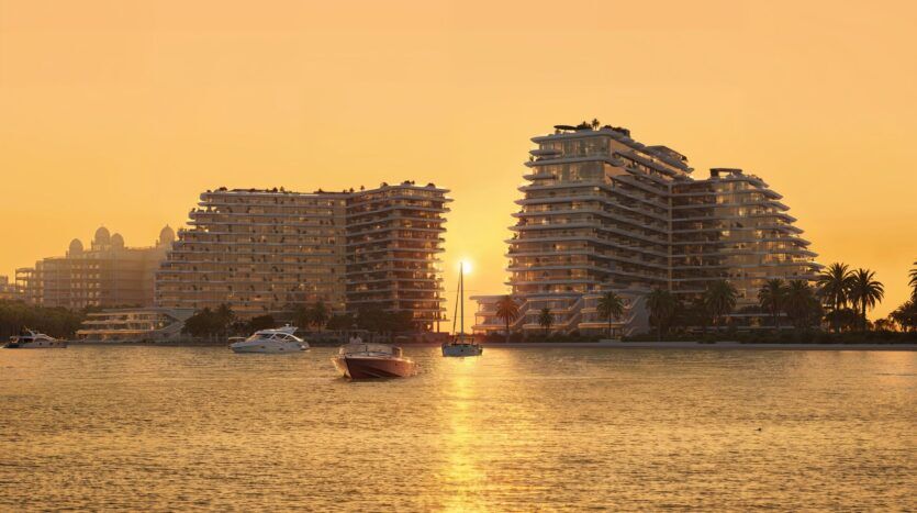 Coucher de soleil sur les bâtiments modernes en terrasses de Bella by Passo, au bord de l'eau, avec des bateaux ancrés dans une eau calme. Le ciel est orangé et des palmiers bordent le rivage, reflétant une atmosphère paisible et luxueuse.