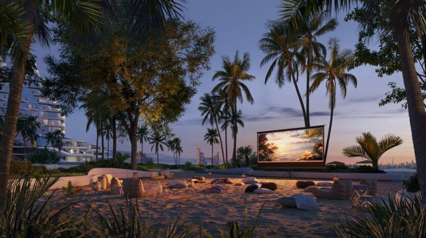 Une installation de cinéma en plein air sur une plage de sable au crépuscule est agrémentée de touches Bella by Passo - des oreillers et des couvertures pour s'asseoir, entourés de palmiers. Un grand écran affiche une scène de coucher de soleil avec des bâtiments modernes en arrière-plan.
