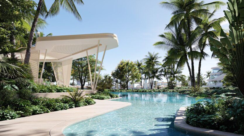 Une piscine claire entourée de plantes vertes luxuriantes et de palmiers, avec le pavillon moderne Bella by Passo sur la gauche, sous un ciel bleu ensoleillé, créant une atmosphère de station balnéaire tropicale.