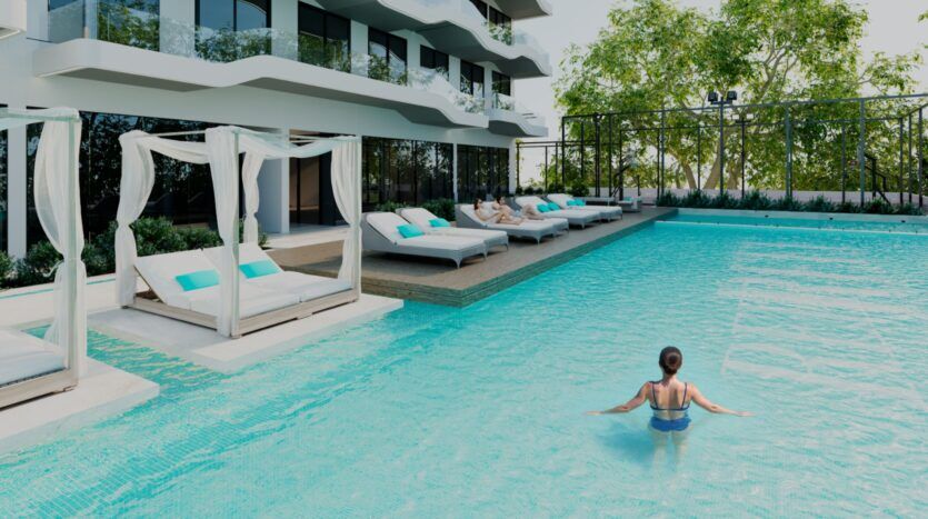 Une femme se tient dans une piscine d'un bleu limpide, à proximité des 996 lits de luxe de Reef, avec des baldaquins blancs et des oreillers turquoise, à l'extérieur d'un bâtiment moderne doté de balcons et entouré d'arbres.