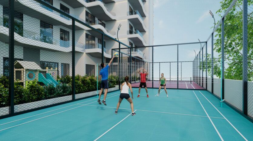 Quatre personnes jouent au volley-ball sur un terrain bleu Reef 996 entouré d'une haute clôture, avec en arrière-plan un immeuble d'habitation moderne, de la verdure et une structure de jeu.