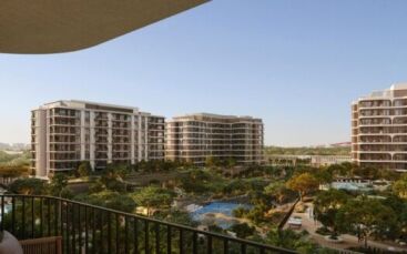 Vue depuis un balcon donnant sur les immeubles d'appartements modernes du complexe « The Canopies by Aldar », entourés d'arbres, d'une piscine et de terrains de sport, sous un ciel bleu limpide.