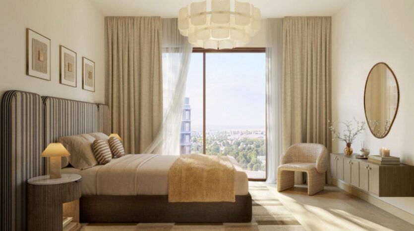 Une chambre moderne de l'établissement « The Canopies by Aldar » comprend un grand lit, une décoration beige, un miroir rond, une fenêtre offrant une vue sur la ville, des rideaux légers, une chaise en bois, des tableaux encadrés et une suspension.