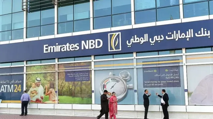 Des gens marchant et conversant devant une succursale bancaire Emirates NBD avec de grandes fenêtres et des publicités pour l'immobilier Dubaï. Un guichet automatique est également visible sur le côté.
