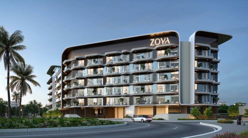 Un immeuble moderne à plusieurs étages, doté de balcons vitrés, d’éléments architecturaux courbes et d’une enseigne « ZOYA », entouré de palmiers et d’espaces verts aménagés au cœur d’un rond-point, sous un ciel d’un bleu limpide : un aperçu séduisant de Mirari Lagoon by Zoya.