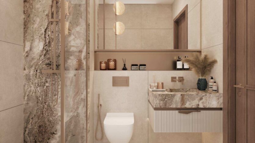 Salle de bains moderne équipée d'un WC suspendu, de plans de travail en marbre, d'un meuble-lavabo suspendu, d'une douche vitrée, de robinetterie dorée, de pots décoratifs et d'herbe de la pampa dans un vase. Les touches de « Mirari Lagoon » de Zoya apportent une note subtile de couleur apaisante aux tons neutres, tandis qu'un éclairage tamisé crée une atmosphère chaleureuse et élégante.