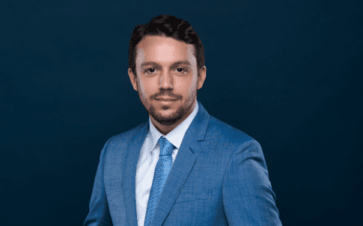 Une photo professionnelle de Guillaume Giroux avec des cheveux bruns courts et une barbe de trois jours claire, vêtu d'un costume bleu et d'une cravate bleue à motifs, sur un fond bleu foncé.