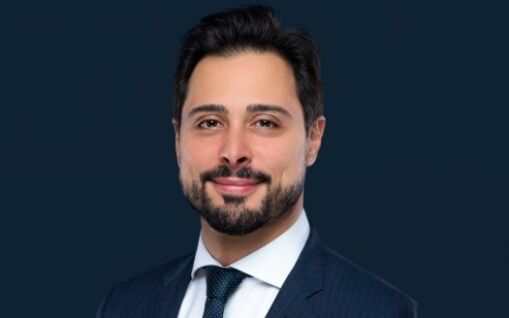Portrait professionnel d'un homme avec un léger sourire, vêtu d'un costume sombre, d'une chemise blanche et d'une cravate bleue, sur un fond bleu foncé. Ses cheveux sont bien coiffés et courts.