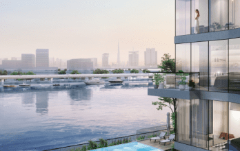 Azura Residences est un complexe d'appartements moderne en bord de mer doté de baies vitrées offrant une vue sur les toits de la ville et la marina. Une personne se tient sur un balcon, surplombant l’eau sereine et les bateaux amarrés en contrebas. Une piscine est visible à un niveau inférieur, renforçant l'atmosphère luxueuse.