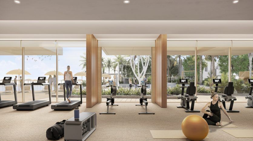 Une salle de sport spacieuse avec des équipements modernes ; une femme qui s'étire sur un tapis et un homme qui court sur un tapis roulant. De grandes fenêtres offrent une vue sur un patio extérieur ensoleillé, soulignant l'attrait de cette villa à Dubaï.